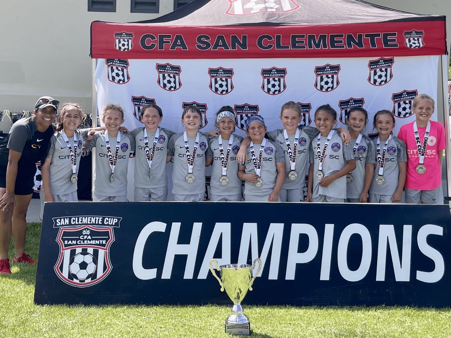 Young girls champions in grey with medals and the trophy under the CFA San Clemente tent