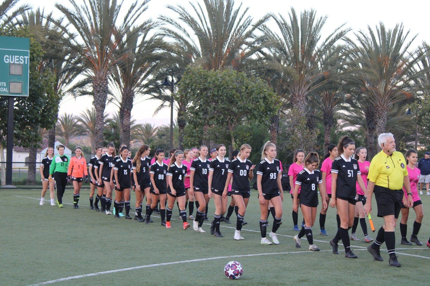 Girls teams lined up on the field before kickoff, with palm trees behind